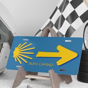 May include: Blue and yellow license plate with a yellow arrow pointing to the right and a scallop shell symbol. The text "BUEN CAMINO" is printed in white below the arrow.