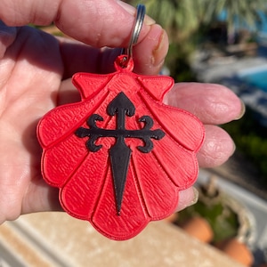 May include: A red plastic keychain shaped like a scallop shell with a black cross and a black arrow in the center.