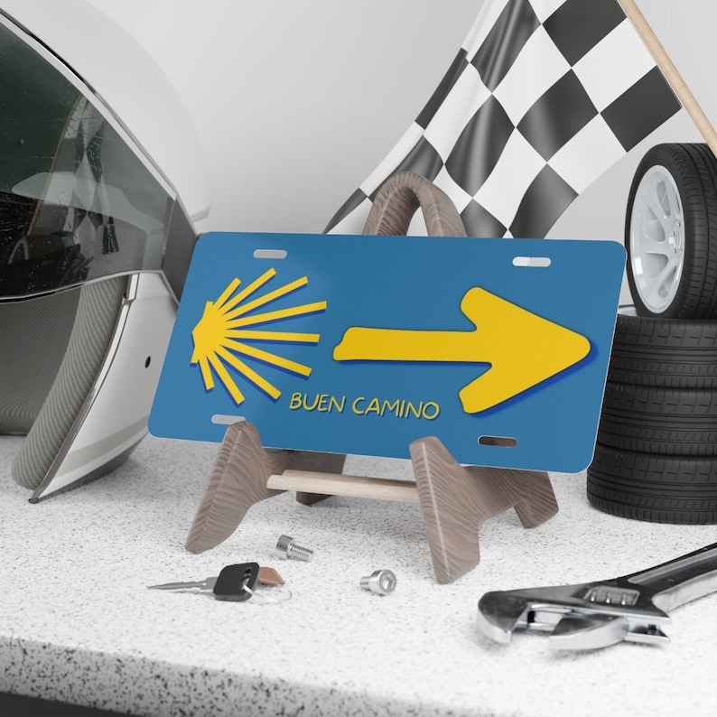 May include: Blue and yellow license plate with a yellow arrow pointing to the right and a scallop shell symbol. The text "BUEN CAMINO" is printed in white below the arrow.