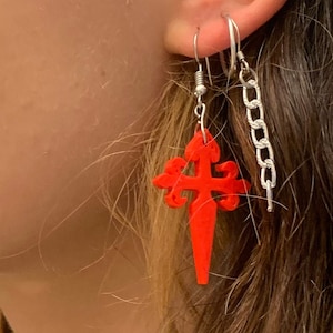 Puede incluir: Un par de pendientes con un colgante de cruz roja y una cadena plateada. La cruz es un diseño estilizado con una parte inferior puntiaguda.