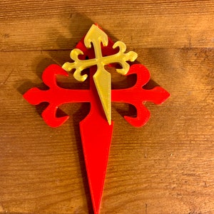Può includere: Una croce rossa e dorata con una punta. La croce è un simbolo tradizionale del Cammino di Santiago.