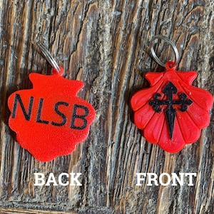 May include: Red plastic keychain with a scallop shell design. The front features a black cross with a sword, and the back has the letters "NLSB" in black.