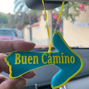 Könnte beinhalten: Ein blauer Pfeil-förmiger Auto-Anhänger in Türkis mit gelber Umrandung und den Worten "Buen Camino" in gelben Buchstaben.