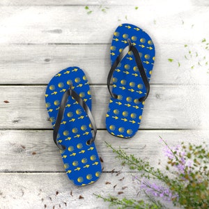May include: A pair of blue flip-flops with a yellow scallop shell pattern. The flip-flops are on a white wooden surface.