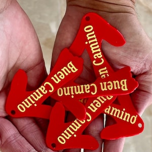 May include: Five red arrow-shaped keychains with the phrase "Buen Camino" written in yellow on each keychain.