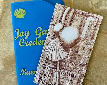 May include: A blue card with a seashell logo and the text "Joy Garden Credencial" in gold lettering. A brown card with a drawing of a seashell and the text "Credencial del Peregrino" is partially visible on top of the blue card.