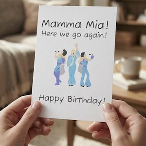 Könnte beinhalten: Eine weiße rechteckige Grußkarte mit dem schwarzen Text Mamma Mia! Here we go again! oben und Happy Birthday! unten. In der Mitte befindet sich eine farbenfrohe Illustration von drei Figuren in blauen Outfits, die Mikrofone halten.