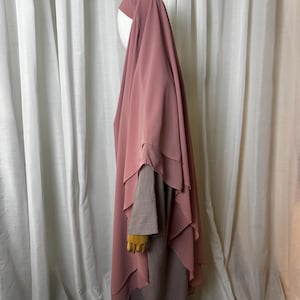 Könnte beinhalten: Ein mehrschichtiges, staubrosafarbenes Hijab und ein passendes Kleid werden auf einer Schaufensterpuppe präsentiert. Das Hijab hat mehrere Schichten aus weichem Stoff, wodurch ein fließender Effekt entsteht. Das Kleid darunter hat lange Ärmel und ein schlichtes Design.