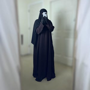 May include: A woman wearing a black abaya and niqab is holding a smartphone in front of her face. She is standing in a room with white walls and a door in the background.