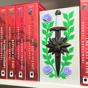 May include: A collection of red-spined books from "The Witcher" series by Andrzej Sapkowski, with a decorative bookend featuring a sword, wolf emblem, and floral accents. The bookend is white, with green, purple, and black details.