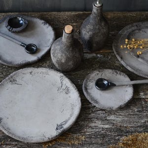 K&ouml;nnte beinhalten: Eine Sammlung handgefertigter, grauer Keramik-Tafelservice und Servierteile. Das Set umfasst Teller, kleine Schalen und Flaschen mit rustikaler, strukturierter Oberfl&auml;che. Ein L&ouml;ffel und ein Aschenbecher sind ebenfalls enthalten. Die Teile sind auf einer verwitterten Holzoberfl&auml;che angeordnet.