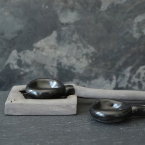 May include: Two black ceramic bowls, one on a gray rectangular base, the other on a gray ceramic handle. The bowls are smooth and glossy.