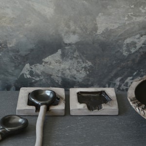 May include: Three black ceramic spoons resting in gray ceramic holders. The spoons are on a dark gray slate surface.