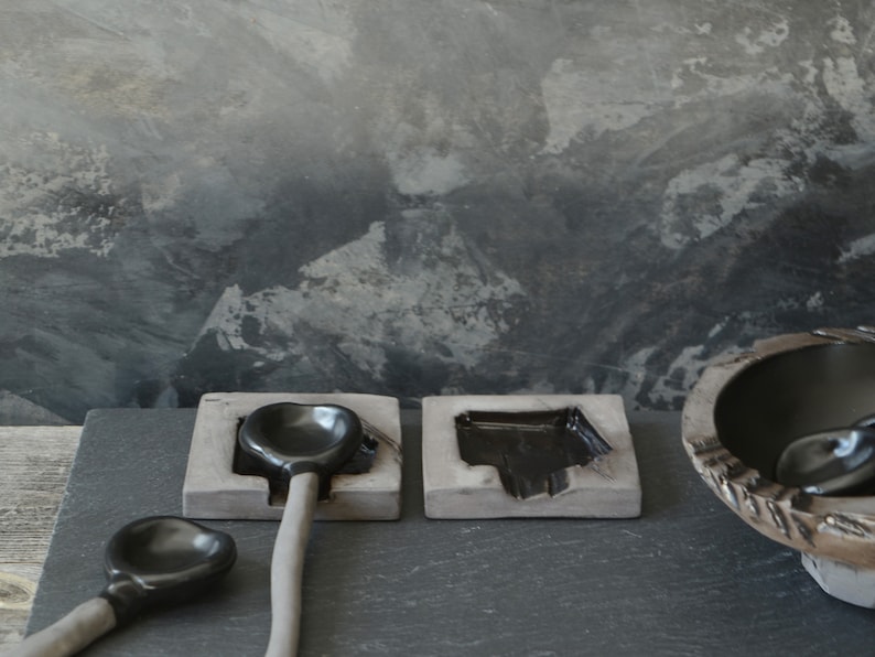 May include: Three black ceramic spoons resting in gray ceramic holders. The spoons are on a dark gray slate surface.