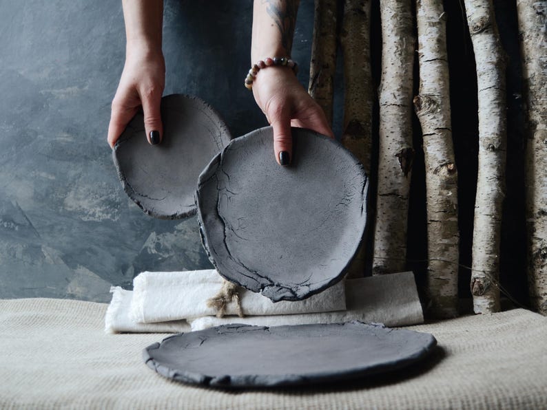 May include: Three gray ceramic plates of different sizes. The plates have a rough, textured surface. The plates are arranged on a light brown linen surface.