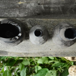 K&ouml;nnte beinhalten: Drei schwarze Keramikvasen in verschiedenen Gr&ouml;&szlig;en und Formen, die auf einer Holzoberfl&auml;che stehen. Die Vasen haben eine raue, strukturierte Oberfl&auml;che.