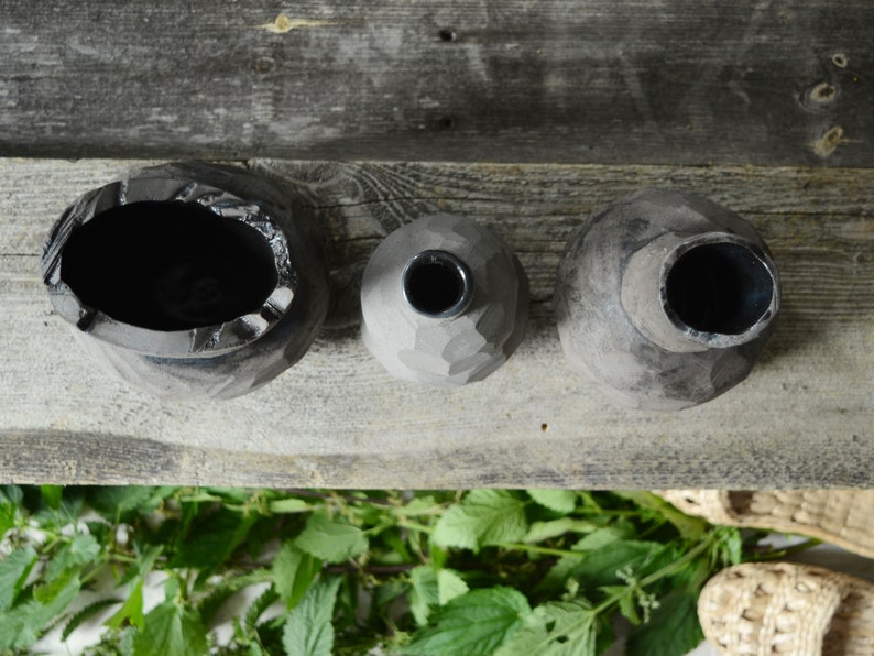 K&ouml;nnte beinhalten: Drei schwarze Keramikvasen in verschiedenen Gr&ouml;&szlig;en und Formen, die auf einer Holzoberfl&auml;che stehen. Die Vasen haben eine raue, strukturierte Oberfl&auml;che.