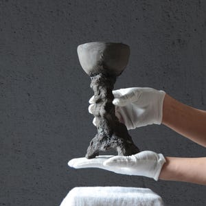 Può includere: Un calice in ceramica grigio scuro, fatto a mano, con uno stelo strutturato e organico e una coppa ampia e poco profonda. Il calice è tenuto da mani guantate su uno sfondo grigio, che mostra il suo design rustico unico.