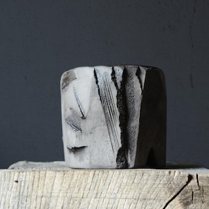 May include: A handcrafted, cylindrical ceramic vase with a textured, grey surface. The vase features carved geometric designs and dark, linear accents. It sits on a weathered wooden block, showcasing a rustic aesthetic.