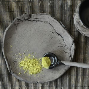 May include: A gray ceramic plate with a rough texture and a small amount of green powder on it. A black ceramic spoon with a green powder in it is resting on the plate.