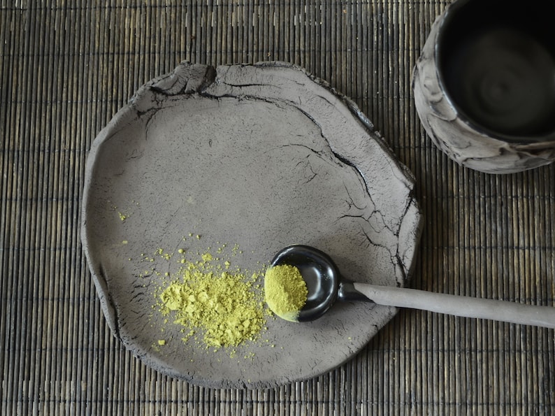 May include: A gray ceramic plate with a rough texture and a small amount of green powder on it. A black ceramic spoon with a green powder in it is resting on the plate.