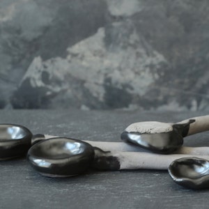 May include: Four black ceramic spoons with rounded bowls and gray handles. The spoons are arranged on a dark gray surface.