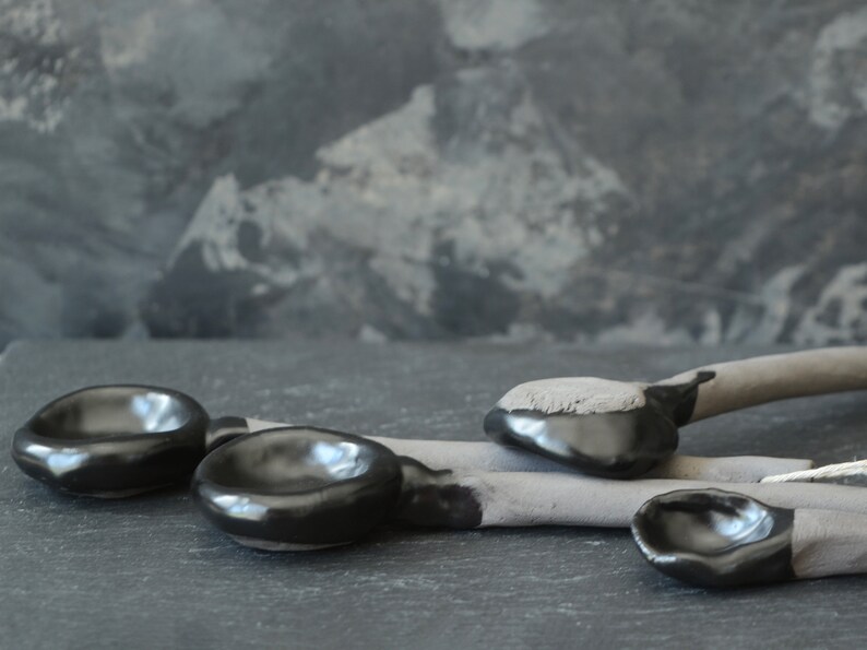 May include: Four black ceramic spoons with rounded bowls and gray handles. The spoons are arranged on a dark gray surface.