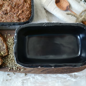 Puede incluir: Un molde para pan negro con base de madera, rodeado de semillas de cáñamo. El molde está vacío, listo para ser usado para hornear.