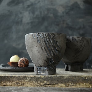 Op de afbeelding: Twee donkergrijze keramische kommen met een getextureerd, ruw oppervlak. De kommen staan op een houten oppervlak met twee chocoladebonbons op een zwart bord op de voorgrond.