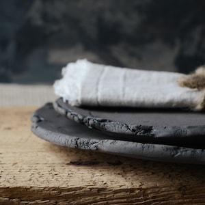 May include: Two black ceramic plates with a rough, textured surface. The plates are stacked on top of each other and are sitting on a wooden surface.