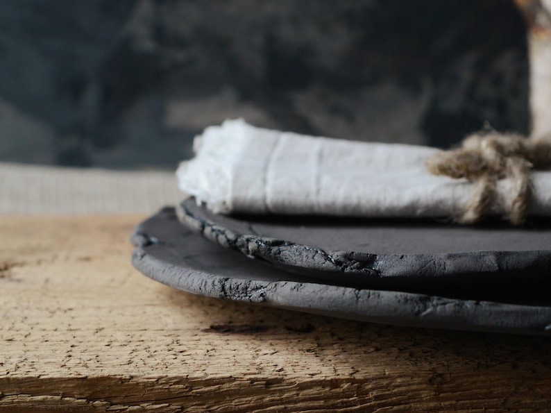 May include: Two black ceramic plates with a rough, textured surface. The plates are stacked on top of each other and are sitting on a wooden surface.