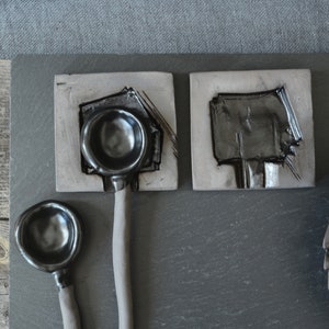 May include: Three black ceramic spoons with rounded bowls, two on square tiles and one on a wooden surface. The spoons have a matte finish and are handmade.