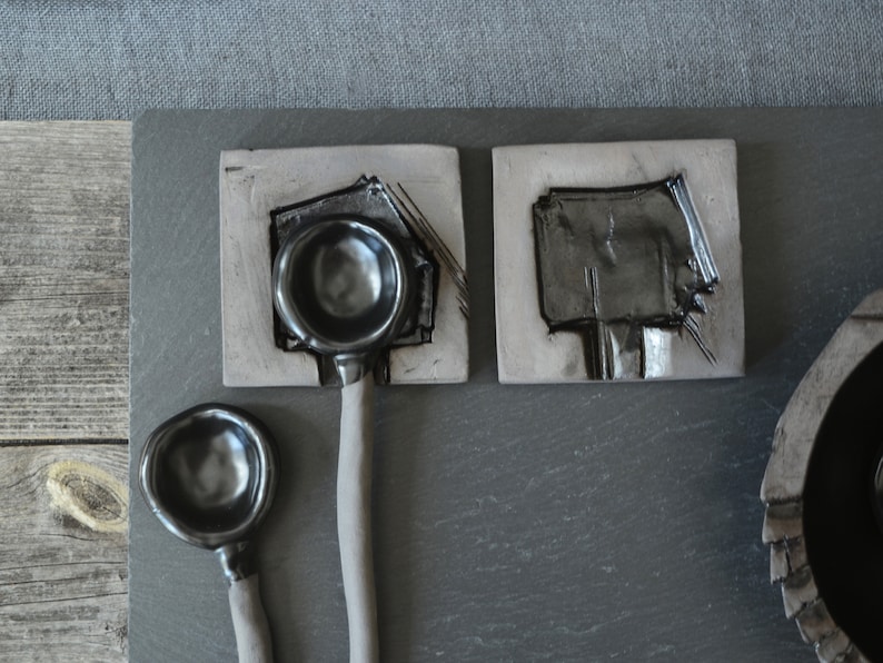 May include: Three black ceramic spoons with rounded bowls, two on square tiles and one on a wooden surface. The spoons have a matte finish and are handmade.
