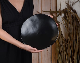 May include: A large, black, round ceramic plate with a slightly curved rim.