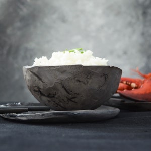May include: A black bowl with a rough, textured surface filled with white rice. The bowl is sitting on a black plate. A red lobster claw is visible in the background.
