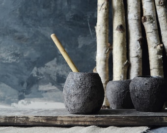May include: A set of three dark gray, round, textured ceramic vessels with a rough, stone-like finish. One vessel contains a light-colored wooden stirring stick. The vessels are arranged on a weathered wooden plank in front of birch logs.