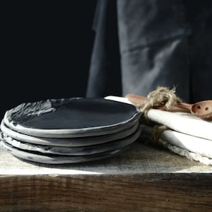 May include: A stack of four black ceramic plates with a rough, textured surface. The plates are arranged on a wooden surface with a white cloth and a wooden spoon in the background.