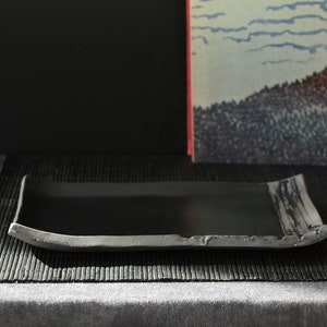 Puede incluir: Una bandeja rectangular negra para servir con un borde ligeramente elevado y una superficie texturizada. La bandeja está sobre una estera negra tejida.