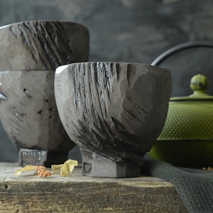 Może przedstawiać: Trzy szare ceramiczne filiżanki do herbaty z fakturowanym, prążkowanym wzorem. Filiżanki są ułożone jedna na drugiej, z największą filiżanką na dole. W tle znajduje się zielony żeliwny czajnik.