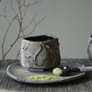 May include: A black ceramic teacup with a textured surface sits on a matching saucer. A small spoon with green powder rests on the saucer.