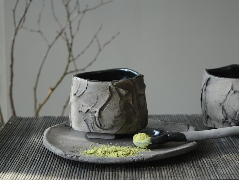 May include: A black ceramic teacup with a textured surface sits on a matching saucer. A small spoon with green powder rests on the saucer.