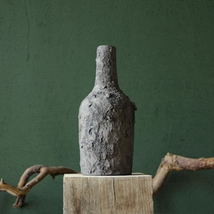 Può includere: Un vaso in ceramica grigia con una superficie testurizzata, che ricorda la corteccia di un albero, si trova su una piattaforma di legno. Il vaso è di fronte a un muro verde.