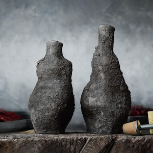 Puede incluir: Dos botellas de cerámica gris oscuro texturizadas con superficies irregulares sobre una superficie de madera envejecida. Tapones y un tapón de botella de metal cerca. El fondo es gris apagado, lo que sugiere un tema rústico o artesanal.