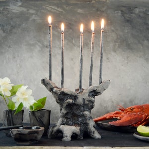 May include: A rustic grey ceramic candelabra with five lit silver candles. The candelabra is on a dark surface with a small vase of white flowers, a small bowl, and a plate with a cooked lobster and lime slices.