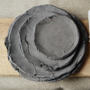 Puede incluir: Un conjunto de tres platos de cerámica gris hechos a mano con una superficie rugosa y texturizada. Los platos están colocados sobre una tabla de madera, con una servilleta de lino blanco atada con hilo a la derecha. Estética rústica y orgánica.