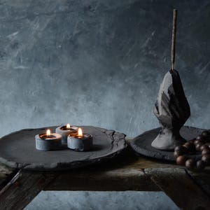May include: A black ceramic incense holder with a pointed top and a stick of incense burning. The holder is on a black ceramic plate with a beaded necklace. Three small candles are burning on a black ceramic plate.