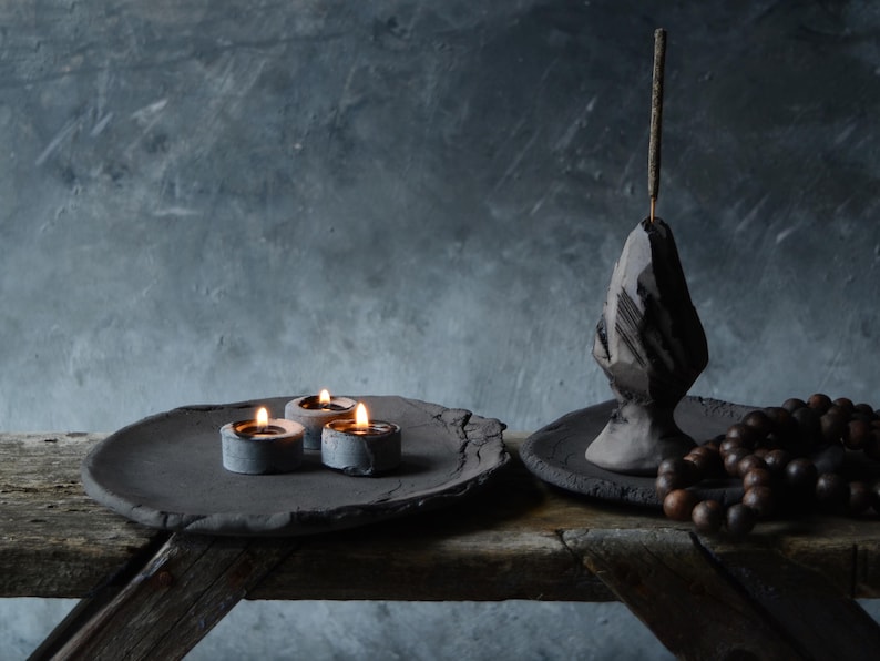May include: A black ceramic incense holder with a pointed top and a stick of incense burning. The holder is on a black ceramic plate with a beaded necklace. Three small candles are burning on a black ceramic plate.