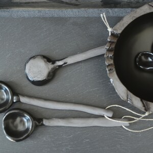 May include: Four black ceramic spoons with gray handles. Two spoons are laying on a gray surface, and two spoons are inside a black ceramic bowl.