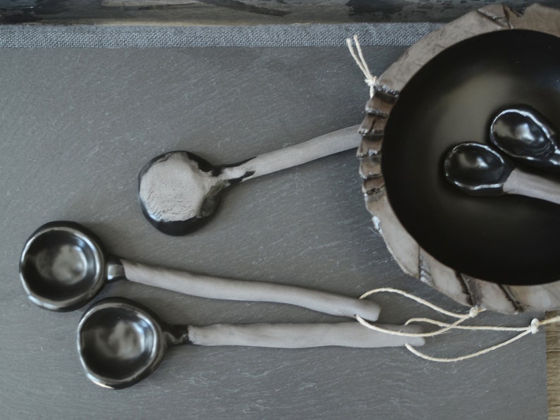 May include: Four black ceramic spoons with gray handles. Two spoons are laying on a gray surface, and two spoons are inside a black ceramic bowl.
