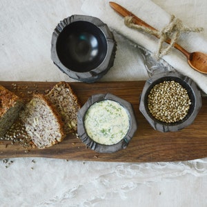 May include: A wooden serving board with three small bowls. One bowl contains a creamy white sauce with green herbs, another bowl contains hemp seeds, and the third bowl is empty. There are also slices of bread on the board.